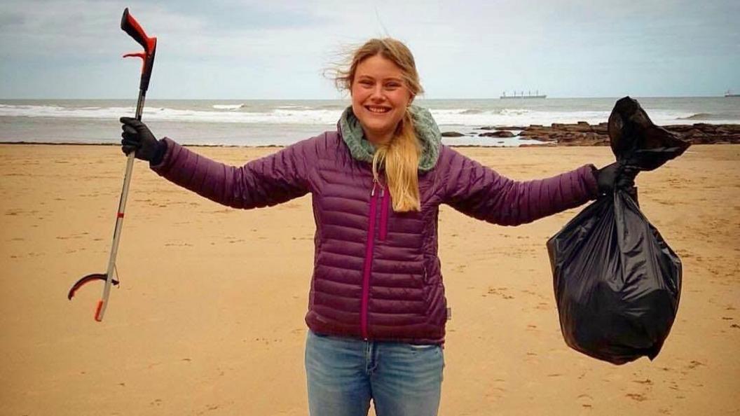 Emily Cunningham standing on a beach with her arms outstretched, holding a bin bag in one hand and a litter picker in the other. She has long blonde hair and is wearing a purple coat, gloves and jeans.