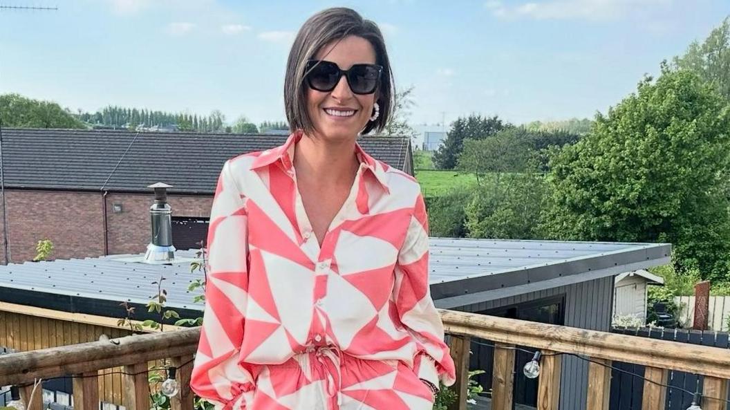 Denise Curran, who has shoulder length dar hair, is wearing sunglasses as she stands on a decking. She is smiling, and wearing a pink and white jacket and matching trousers. A number of buildings can be seen in the background