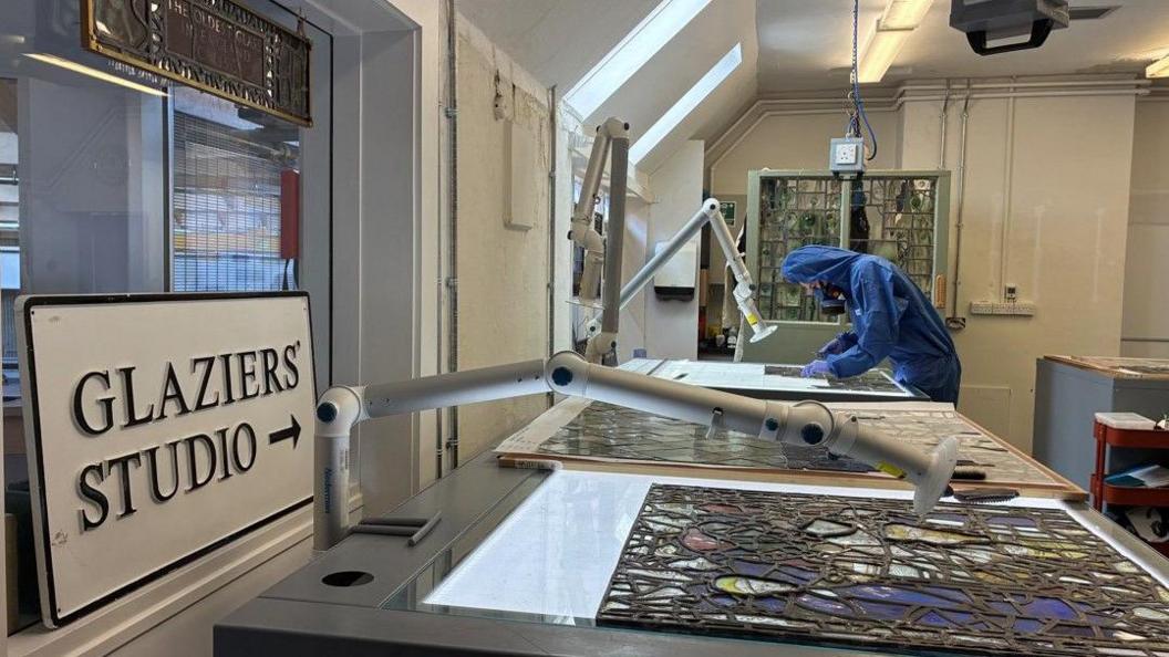 A room with a sign saying Glaziers' Studio on the foreground. A person in a blue protective top with a hood leans over a lit up table with a panel of stained glass on top of it.