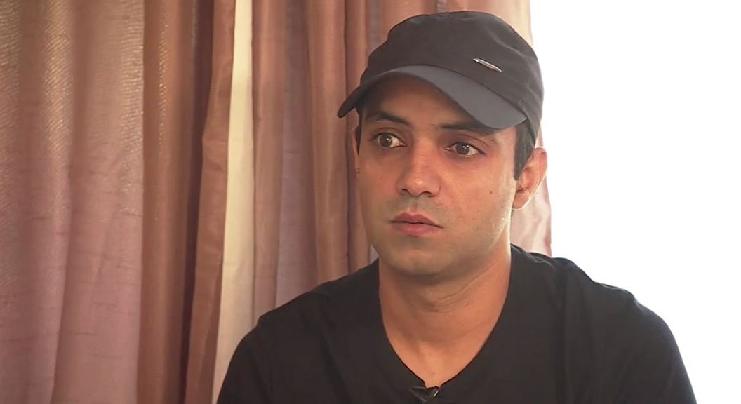 Nabeel Ahmad looking straight into the camera, wearing a black t-shirt and a black baseball cap with pink curtains behind him.