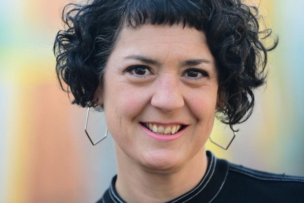 Laura Murphy, a young woman with black curly hair and octagonal silver earrings, smiles at the camera against a neutral, light blue background