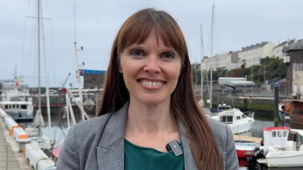 Joney Faragher has long brown straight hair and a fringe. She is smiling, and wearing a green top under a grey suit jacket. She is standing in front of the quay in Douglas, with many boats behind her under grey skies.
