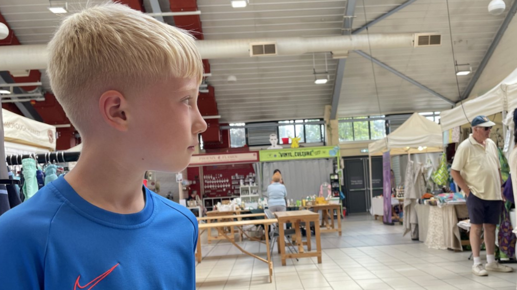 Blonde boy in blue t shirt, looking off to the side at an indoor market
