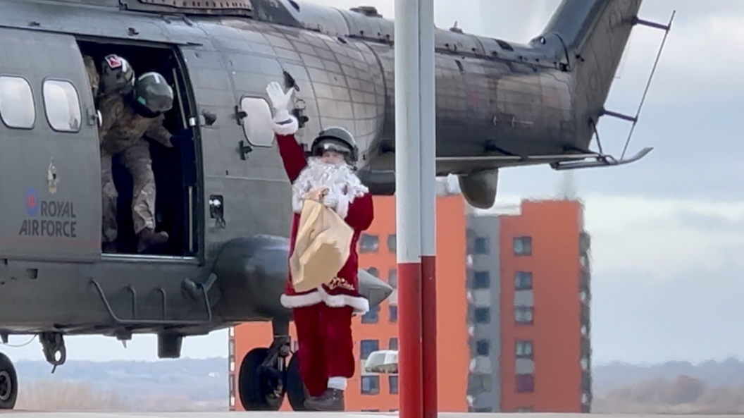 From the top of a building a man dressed as Santa waves to unseen people after stepping off a large RAF helicopter. He is holding a brown sack full of presents and a the top of an apartment building can be seen in the background