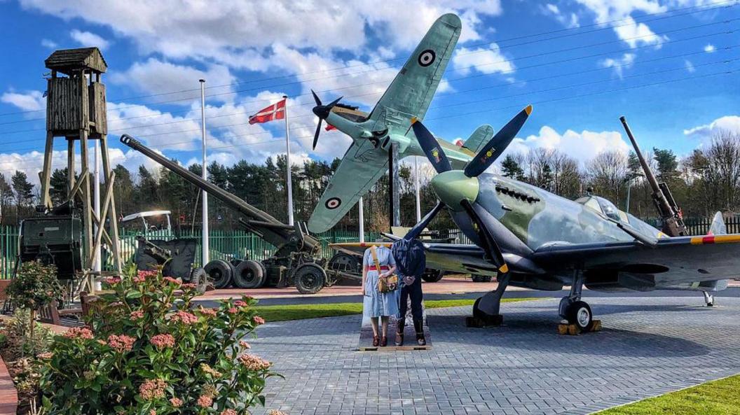 Two World War War 2 in the middle of the picture with mannequins of two military personnel standing in front of the planes on a brick laid driveway. Other WW2 military equipment, such as guns and a wooden look out tower behind them.  