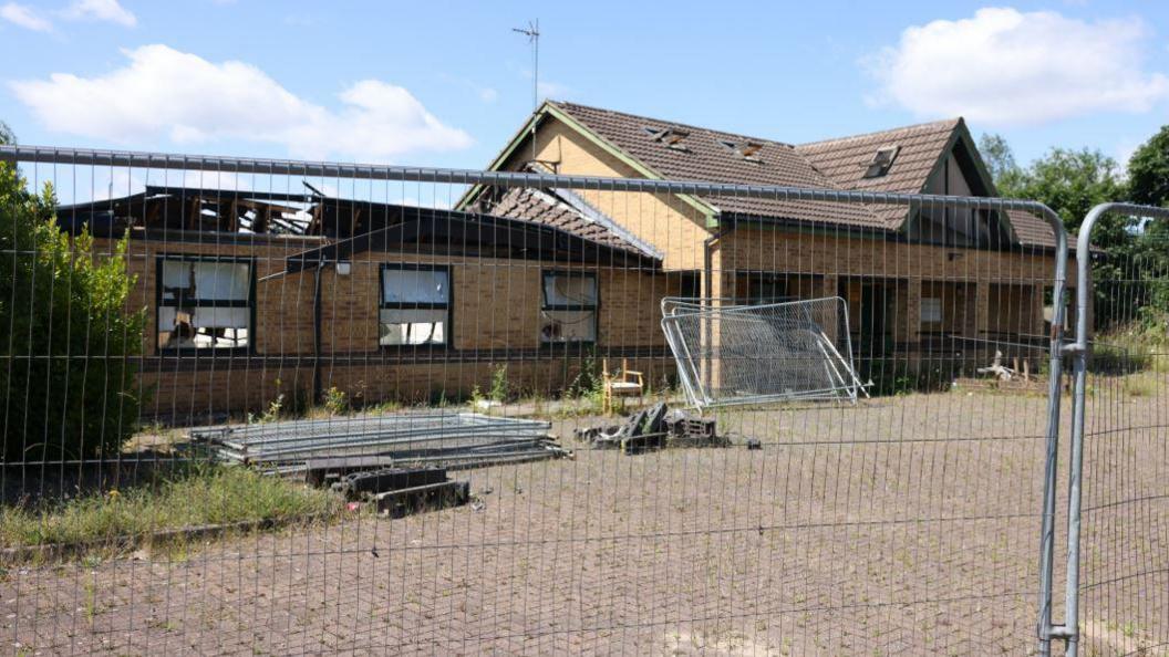 Stanton Lodge Care Home. The building is surrounded by a metal fence. The building is made from light-coloured bricks. Its windows are smashed and part of its roof has collapsed in.