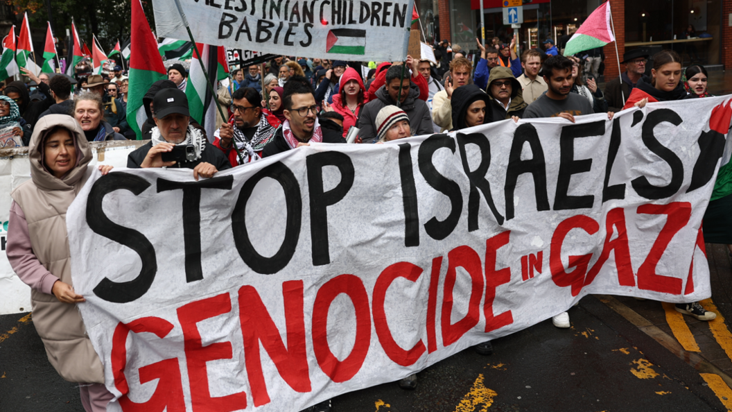 A large crowd, many holding Palestinian flags, marching along a street. At the front several people hold a large white banner with the words Stop Israel's Genocide in Gaza on it. 