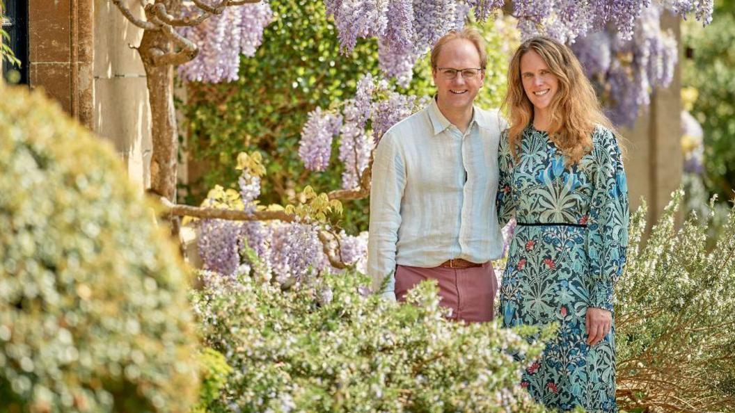 William and his wife Marianne Cartwright-Hignett standing a garden on part of the grounds of the manor. They are surrounded by Wisteria and rosemary.