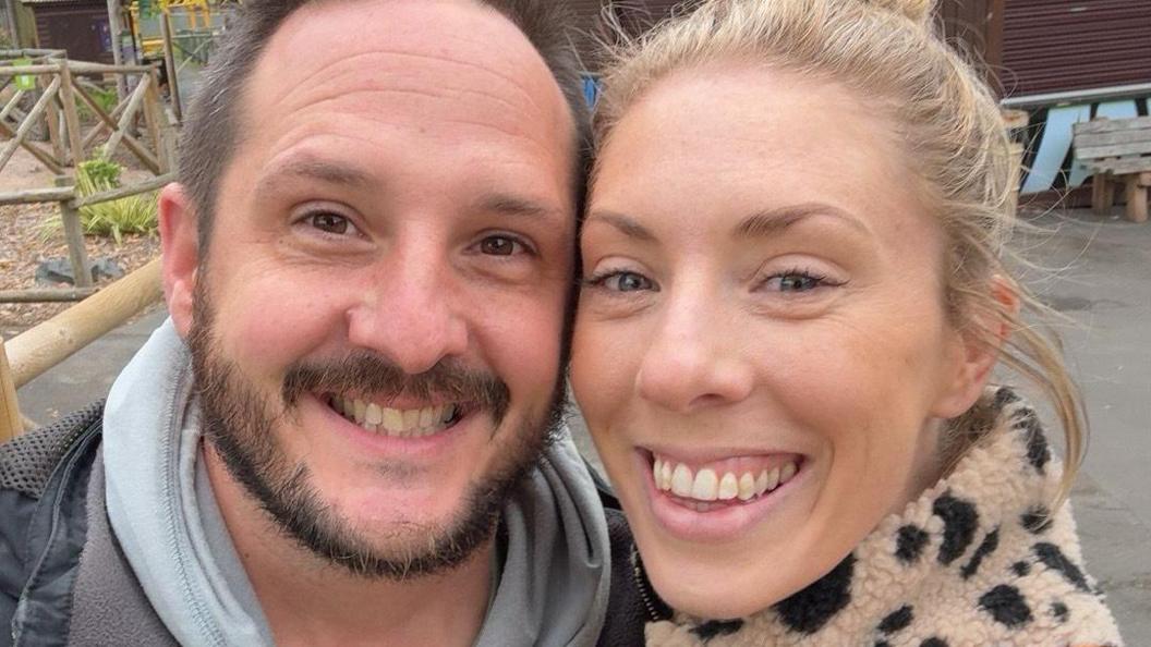 A selfie of Andy and Daisy. Andy is wearing a grey hoodie and dark coat, and has dark hair and a dark beard. Daisy is wearing an animal-print fleece and has light blonde hair pulled back in a bun. Both are smiling widely at the camera. 