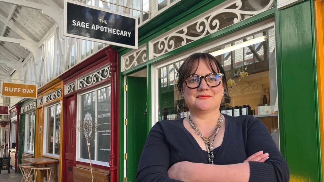 Abi Jackson wearing glasses, necklaces and a blue top, in front of her shop, The Sage Apothecary in Bideford Pannier Market