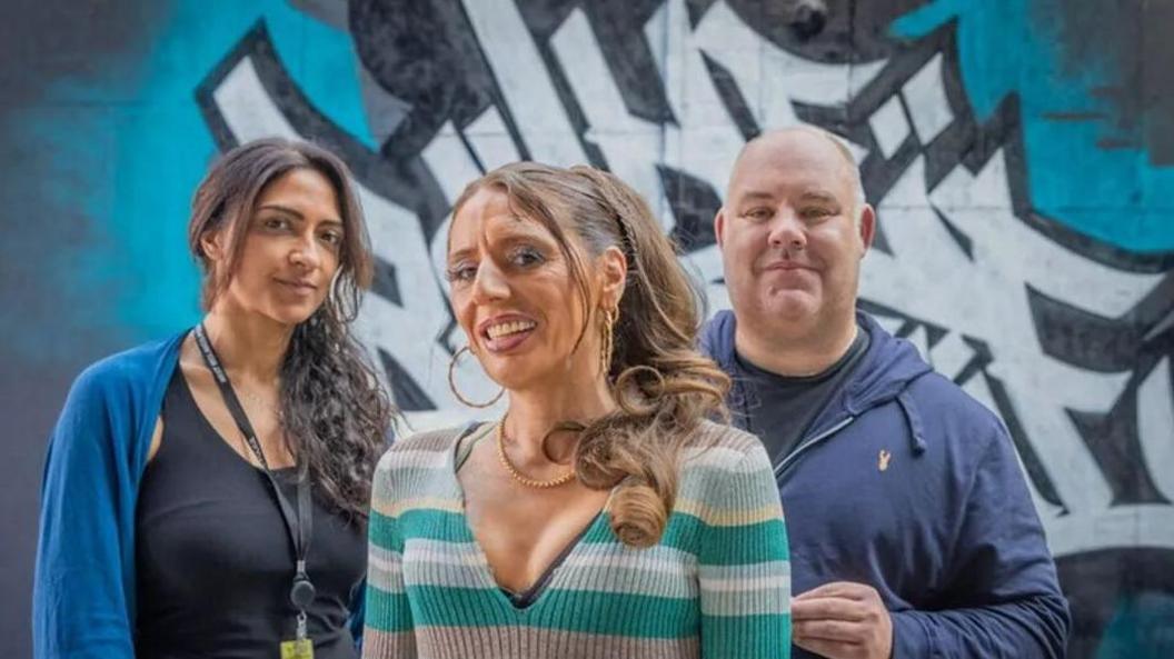 Keeley Knowles is pictured in the middle of two police officers. She is smiling and has her hair in a ponytail and is wearing a blue and grey stripe V-neck long sleeve top. The female officer on her left has curly shoulder length black hair, is wearing a blue cardigan and black tank top. The male officer is bald and wears a blue hoodie on top of a black crew top shirt.
