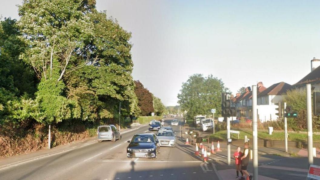 Holme Lacy Road in Hereford in the sunshine at a traffic junction with cars queuing and shops and trees either side of the road. People can be seen on the pavement.