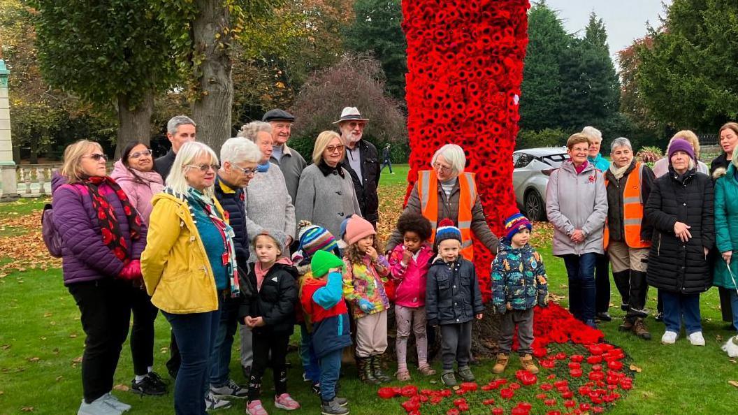 A crowd gathered to pose near a tree adorned with knitted poppies around its trunk