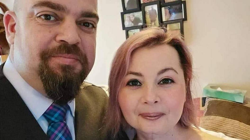 Laura, who has pinkish shoulder-length hair, stands next to her husband, who is wearing a colourful tie and jacket