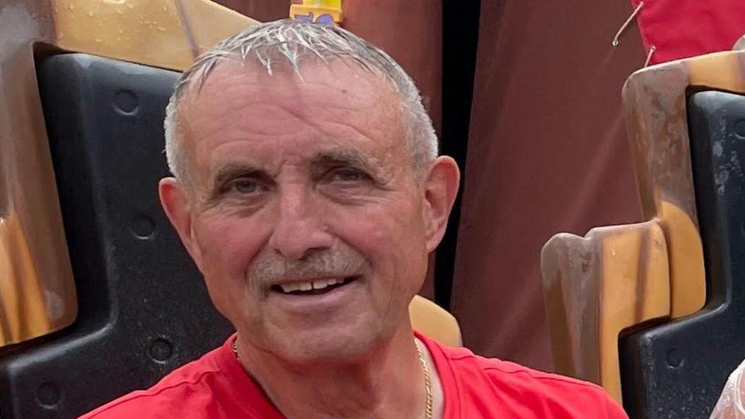 An older man with short grey hair, wearing a red t-shirt, looks into the camera and smiles.