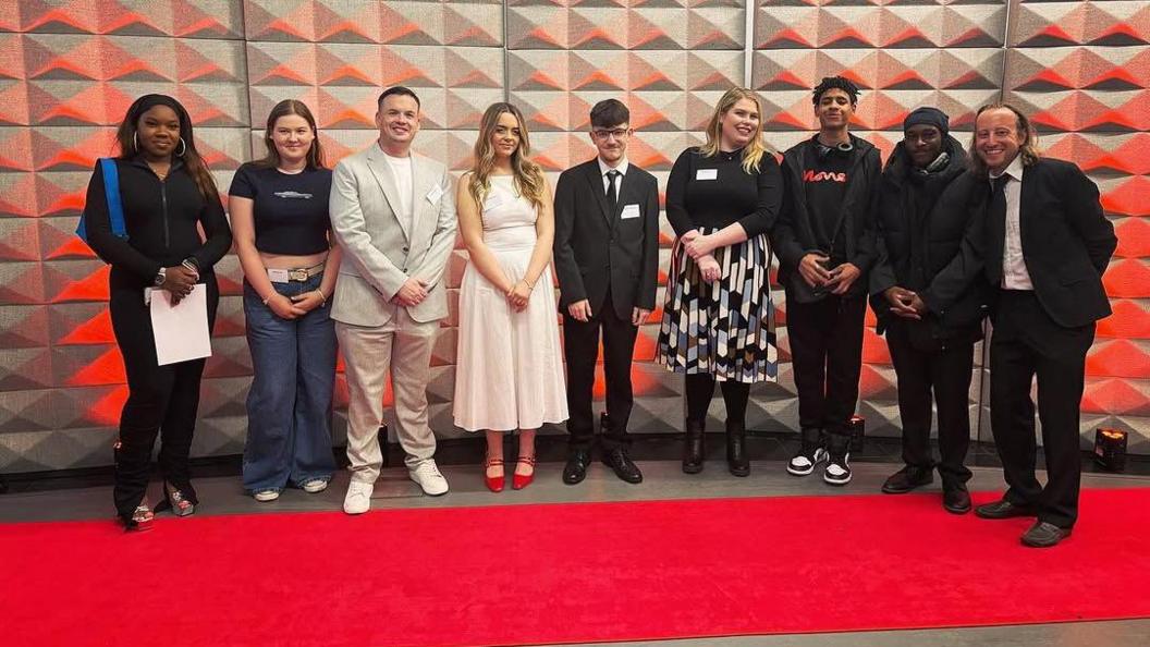 A line up of college students dressed in black tie posing for a picture against a grey 3D backdrop, standing on a red carpet.