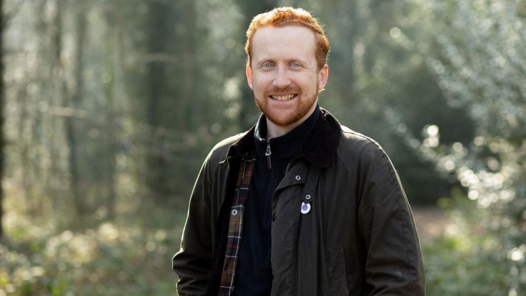 A man with ginger hair and a ginger beard stood in a forest smiling for the camera. He is wearing a dark jacket.