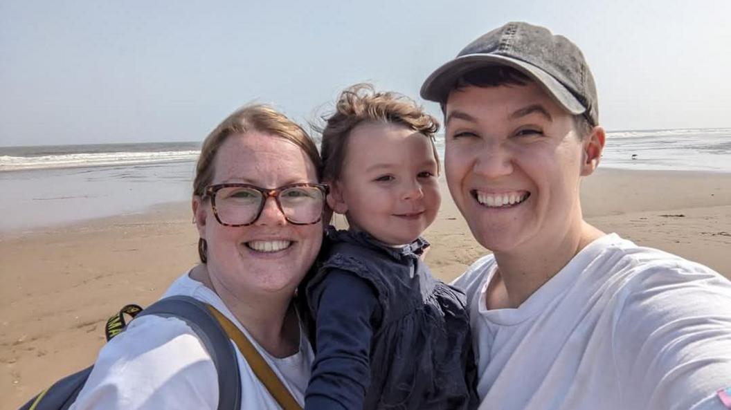 Ellie Bright (left) and Ali Bright (right) holding their three-year-old daughter smiling at the camera. A beach and the sea are in the background.