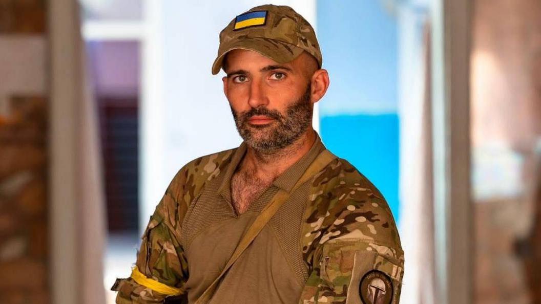 Daniel Burke wears green military fatigues, including a shirt and cap bearing the Ukranian flag. He is sat indoors, outside the entrance to a doorway, with daylight streaming in behind him. 