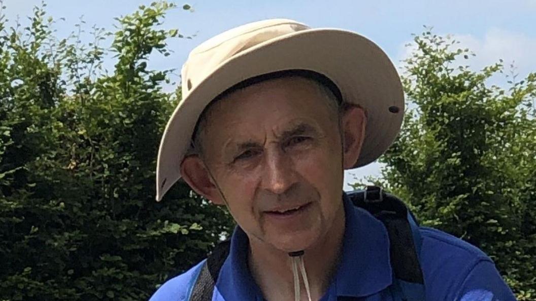 A headshot of Prof Saunders wearing a khaki wide-brimmed hat and a blue polo shirt with a rucksack. There are trees and blue skies in the background. 