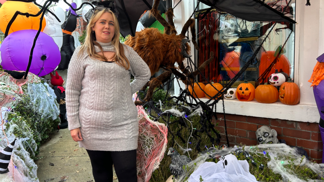 Claire Cox, a woman with long blonde hair in a grey dress, stands outside her house covered in Halloween decorations