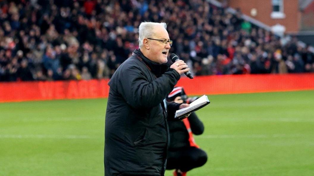 A side shot of Alan Long wearing a long black coat and glasses. He is holding a notebook in his left hand and is holding a black microphone up to his mouth with his right hand. He is stood on a football pitch. The crowd is blurred in the stadium in the background. 