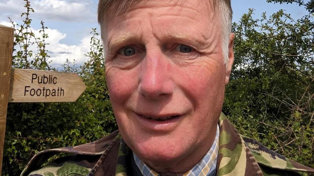A selfie of a man with short grey and brown hair. He is smiling at the camera. He is wearing a cargo jacket and blue check shirt. Behind him is a country lane hedge and public footpath sign.
