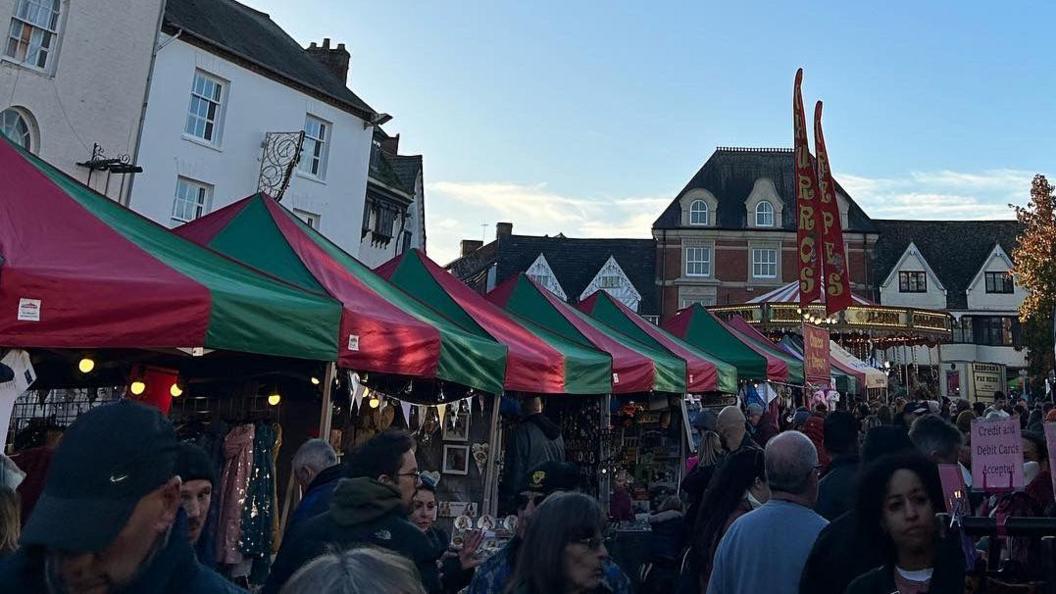 Market stalls are either side of a walkway. They all have red and green roofs. Hundreds of people are walking in the middle. At the end of the walkway is a carousel.