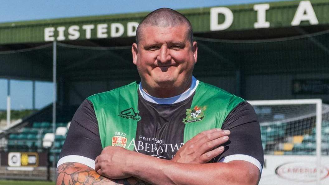 Gareth Ward, who has a buzz cut, stood in an Aberystwyth Town Football Club top which is black with green stripes down it. He is stood in the football grounds with his arms folded in front of him.  