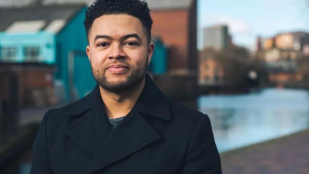 A man in a black tailored coat standing in front of a stretch of the canal in Birmingham city centre