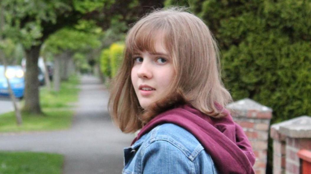 Alex looks back towards the camera. Alex has shoulder length mousy brown hair, with a fringe and wears a blue denim jacket and marron coloured hoodie underneath. In the background is a pathway lined by trees along a residential road.