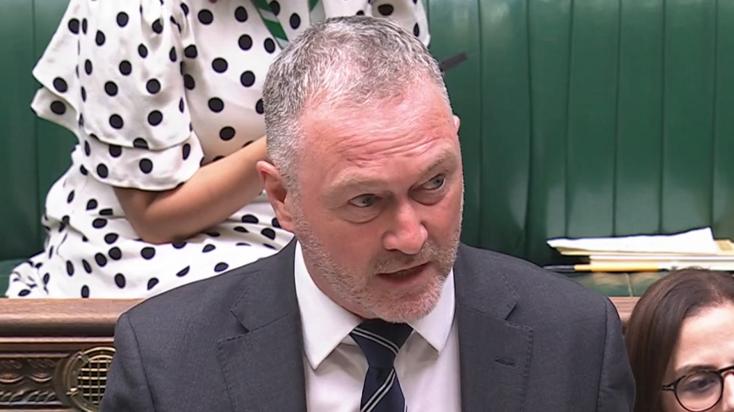 Steve Reed is standing in the House of Commons and is speaking. He has short grey hair and is wearing a white shirt, navy tie and a grey suit jacket.