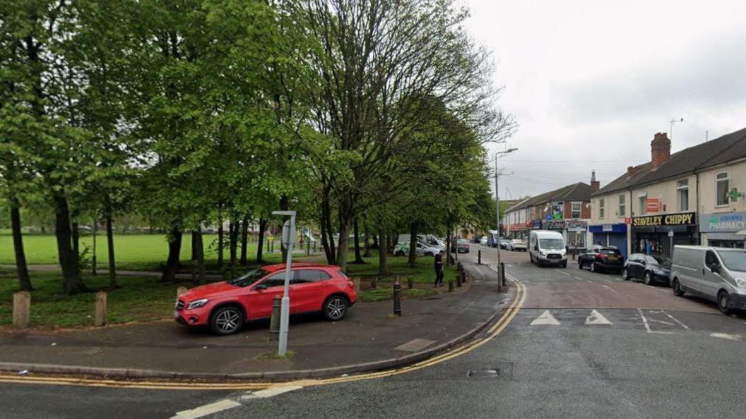 A red car is parked on the pavement adjacent to a park. Trees can be seen on one side of the road, with shops on the other side. A number of vehicles can be seen parked on the kerbside.