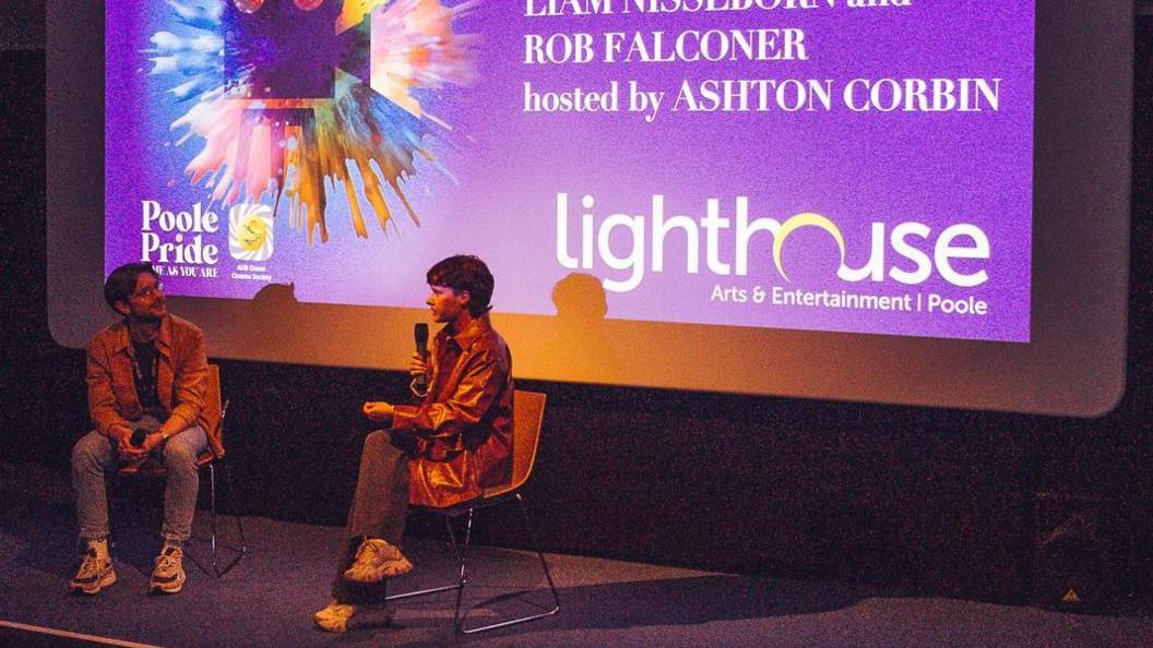 Two white young men sit down on a stage, one of them is director Liam Nissenborn, who is talking into a microphone 