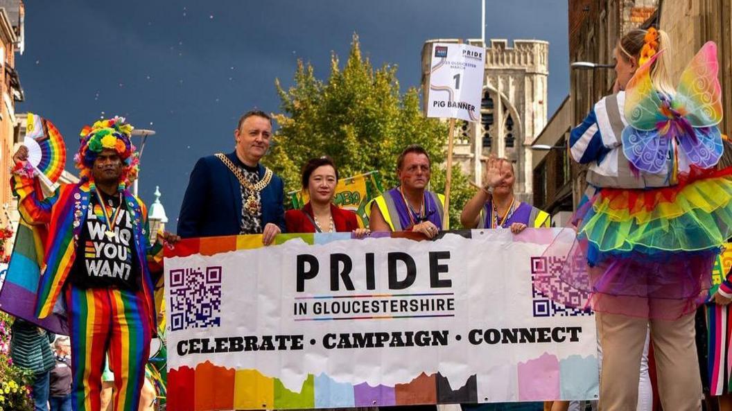 A group of people take part in a Pride parade in Gloucestershire holding a rainbow-coloured banner reading “PRIDE IN GLOUCESTERSHIRE” with the words “CELEBRATE • CAMPAIGN • CONNECT” beneath. The banner features QR codes at each end. People wear vibrant outfits, including a rainbow tutu and a top with the phrase “LOVE WHO YOU WANT”. A historic building and trees are visible in the background.