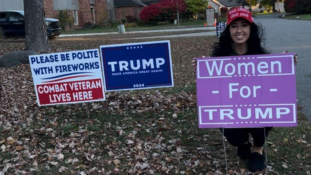 Amanda Sue Mathis wears a red Make America Great Again hat while crouching behind a purple Women for Trump sign in her yard, which is full of autumn leaves. Other signs include a navy Trump campaign sign and a sign that says: "Please be polite with fireworks. Combat veteran lives here."