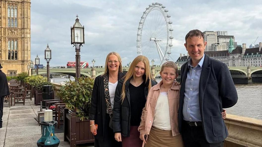 Millie, standing outside at the House of Commons, with the London Eye behind her father, mum and sister. They are all smiling and looking at the camera.