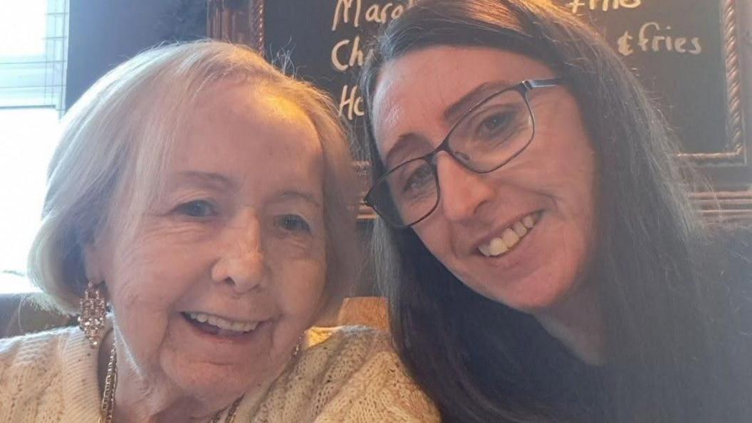 Elizabeth Anne Shipton (left) and Tracy Faram Shipton (right) are photographed taking a picture together using a front facing camera. Elizabeth has short light hair and is wearing a cream knitted jumper. She is smiling and looking at the camera. Tracy has long dark hair and is wearing a black top and glasses. She is also smiling and looking at the camera.