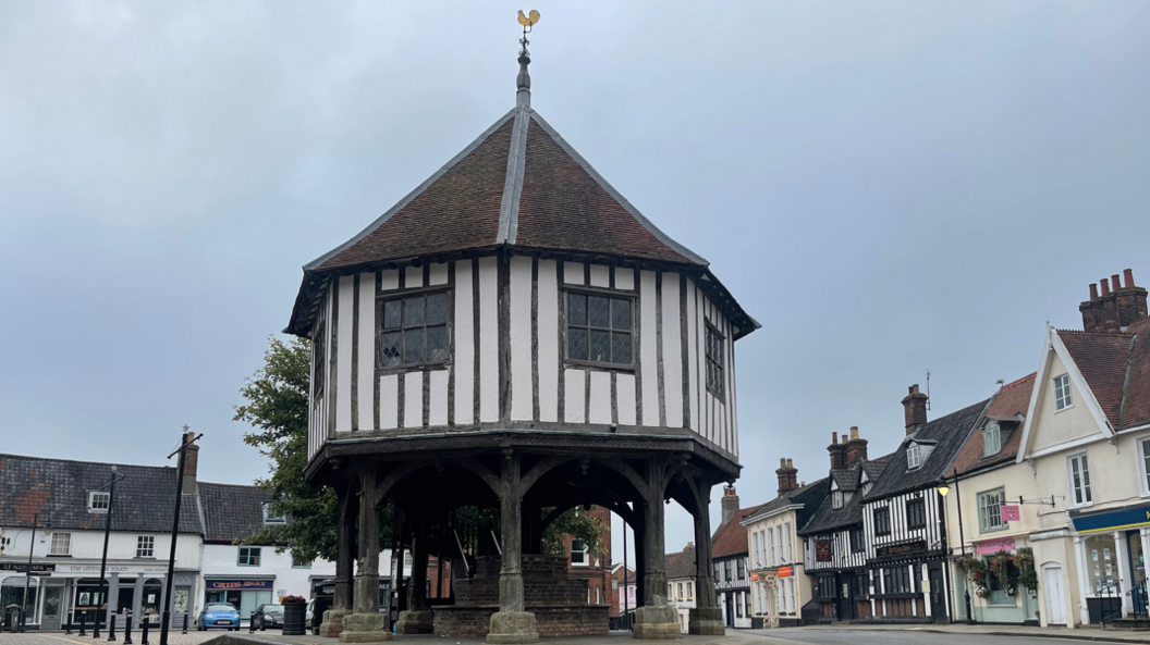 Market Cross in Wymondham, a timber-framed octagonal building supported by posts with other historic buildings containing shops, cafes and pubs behind it