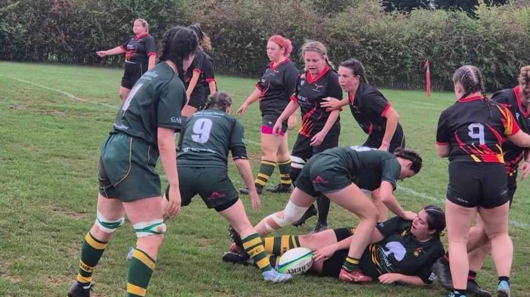 Female teams in green and yellow as well as black and red playing a rugby match.