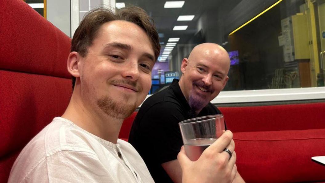 Ali Churchill and Phil Churchill. They are sat indoors on a red sofa. Ali is closest to the camera, smiling at it, and has short dark brown hair and facial hair. Phil, his dad, is sitting beside him. He is bald and has a purple dyed beard and is smiling at his son.