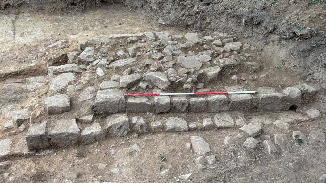 The remains of a wall uncovered from the ground. A red and white stick is on top of some bricks showing the size of the wall.