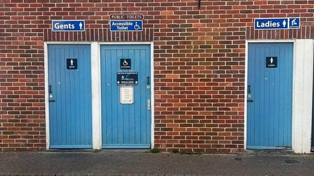 The entrance of a public toilet facility. It's a brick wall with three light blue doors - a disabled toilet, a men's toilet and a women's toilet.