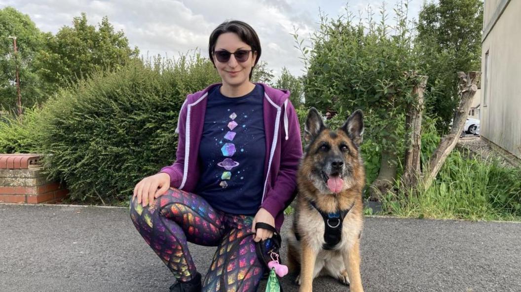 A woman in sunglasses, a purple sweater, dark tee-shirt and shiny scale-pattern leggings kneels next to a German Shepherd dog on a path in front of a hedge. 