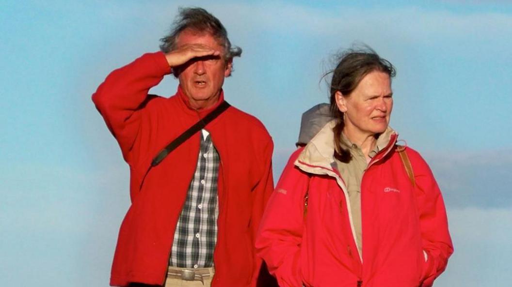 Miranda Mugford and her husband Mike standing next to each other. The are both wearing red outdoor-type coats. Mike has his right hand above his eyes, to avoid sun glare.