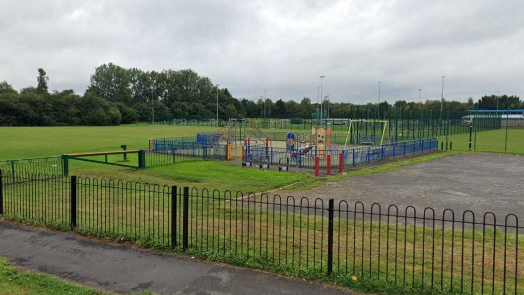 A children's play park surrounded by fencing, with colourful play equipment and a large playing field to the side of it.
