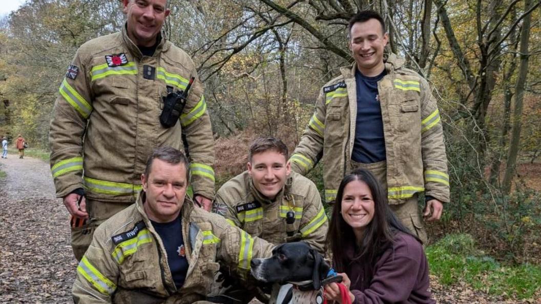 Four firefighters, a dog and a woman in a wooded area. Two firemen are standing, and two are crouching next to a black and white dog and its owner, a woman wearing a purple jacket