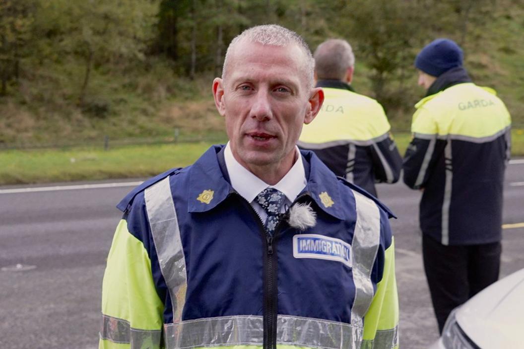 Det Ch Supt Aidan Minnock, with short grey hair and wearing a blue and yellow fluorescent Garda uniform with an "immigration" label, faces the camera while two other Garda officers stand behind him on the road 