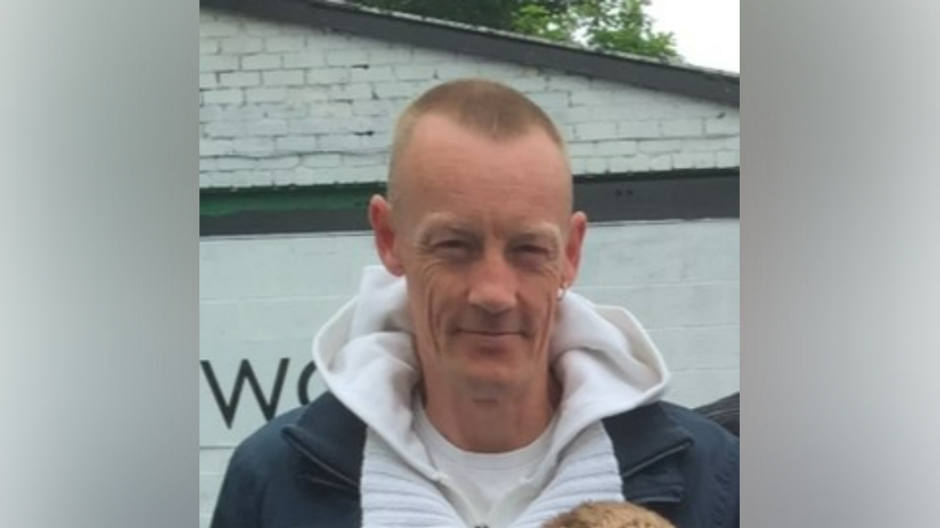 A man with short hair and one gold earring, wearing a white hoodie and navy coat, smiles at the camera.