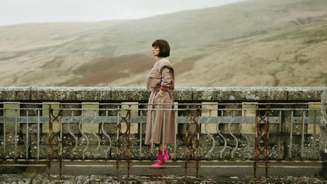 A screengrab from Wild Animal. It shows a woman with a short brown bob wearing a beige outfit and bright pink boots standing on a bridge footpath